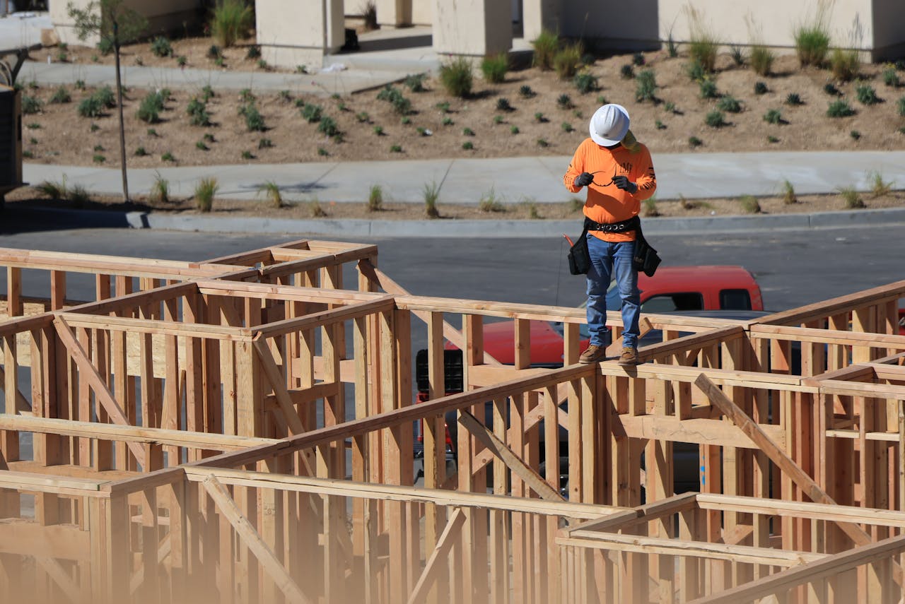 A construction worker on a wooden frame checks a device at a building site.